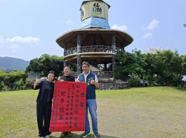 臺南之光!仙湖休閒農場勇奪第19屆世界農業 旅遊大賽「文化與遺產保護類別」世界冠軍 Three people pose on a grassy field, holding a red banner with Chinese characters, in front of a circular wooden pavilion under a blue sky.