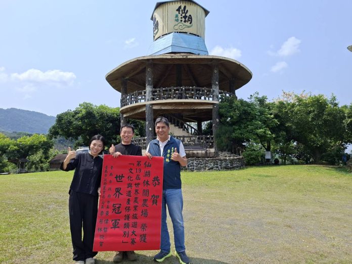 Three people pose on a grassy field, holding a red banner with Chinese characters, in front of a circular wooden pavilion under a blue sky.
