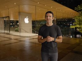 Man in a black t-shirt and jeans stands outside an Apple Store at night, hands clasped in front.