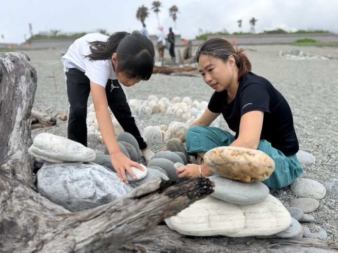 Mother and daughter kneeling on a pebbled beach, stacking smooth stones into a small rock formation.